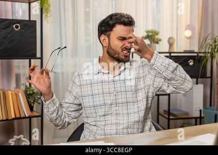 Überarbeiteter junger indischer Geschäftsmann, der an Augenschmerzen und Kopfschmerzen leidet und am Laptop arbeitet, der am Schreibtisch im Heimbüro sitzt. Ein müder hispanischer Freelancer zieht Brillen und Massagen aus, reibt sich die Nasenbrücke Stockfoto