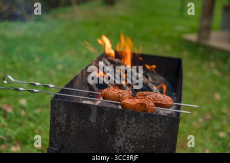 Braten von Würstchen auf Spießen über offener Flamme im rustikalen Metallgrill während des Grillens im Hinterhof. Stockfoto