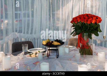 Eine intime romantische Dinnerszene bietet einen großen Strauß roter Rosen, eine Obstplatte und zündete Kerzen auf einem Tisch mit weißem Stoff. Ideal für Stockfoto