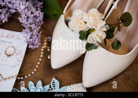 Elegante Hochzeitsvorbereitungsszene mit Elfenbeinabsätzen mit weißen Blumen, lilafarbenen Zweigen, einer Perlenkette und einer Hochzeitseinladung Stockfoto