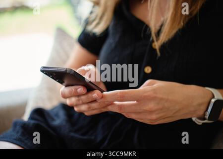 Nahaufnahme einer Frau mit Smartphone, während sie auf der Couch sitzt. Frau mit schwarzem Kleid und Smartwatch, die zu Hause auf dem Sofa sitzt und das Mobiltelefon benutzt. M Stockfoto