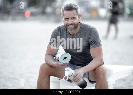 Sport-Fitness-Mann, der draußen auf einer Bank ruht. Reifes Fitnesssportmodell, das nach dem Training auf einer Bank sitzt. Senior Sports man macht danach eine Pause Stockfoto