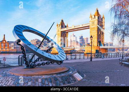 An der Themse steht eine große Sonnenuhr mit der Tower Bridge im Hintergrund. Die Sonne wirft Schatten auf die Sonnenuhr und markiert die Zeit, während Touristen die Aussicht genießen. Stockfoto