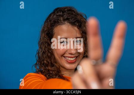 Fröhliche junge Frau, die Friedenszeichen mit Fingern auf leuchtendem blauem Hintergrund bewegt Stockfoto