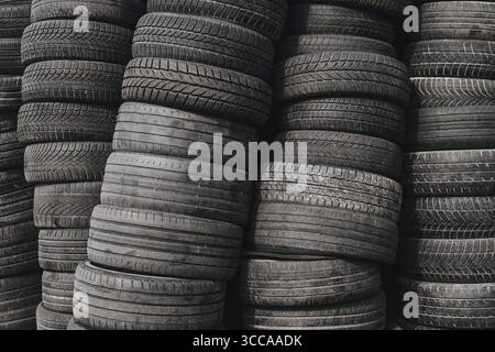 Gestapelte Altreifen in verschiedenen Laufflächenmustern, in hohen Stapeln angeordnet, in Schwarzweiß fotografiert für einen rauen industriellen Textureffekt. Selektiv Stockfoto
