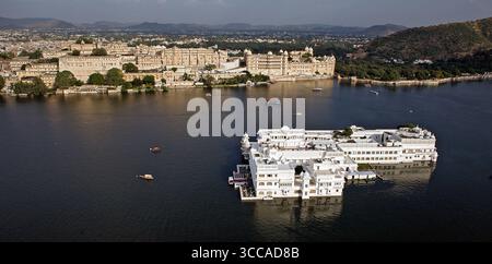 Aus der Vogelperspektive auf den glitzernden Pichola-See schlängelt sich der unberührte weiße Taj Lake Palace und der Stadtpalast vor dem Hintergrund der Skyline von Udaipur, Udaipur, Rajasthan, Indien. Stockfoto