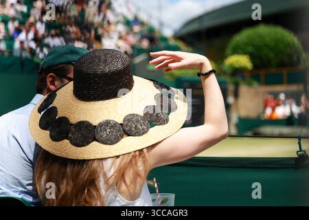 Eine junge Frau mit einem großen Strohhut, die auf einem der Außenplätze bei den Wimbledon Championships 2025 sitzt, London, England, Großbritannien. Stockfoto