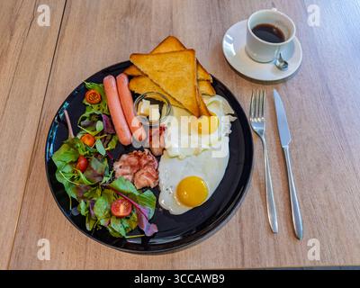 Eine herzhafte und komplette Frühstücksplatte mit Kaffee, mit sonnigen Eiern, Würstchen, knusprigem Speck und Toast. Ein appetitlicher und perfekter Kaffee Stockfoto