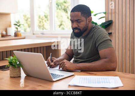 Millennial afroamerikanischer Mann, der sich während eines Webinars oder eines Geschäftstreffens Notizen macht, Laptop benutzt, online arbeitet oder studiert, zuhause am Schreibtisch sitzt Stockfoto