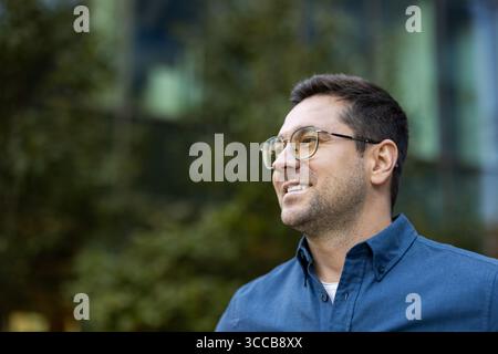 Ein fröhlicher Mann mit Brille lächelt natürlich draußen, trägt ein blaues Hemd, mit verschwommenem grünem Laub im Hintergrund. Stockfoto