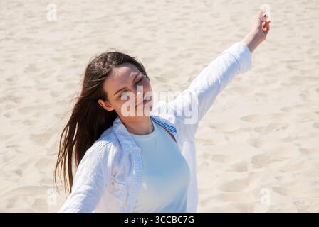 Junge Frau mit langen braunen Haaren, die am Sandstrand steht, mit geschlossenen Augen und erhobenen Armen, genießt das Sonnenlicht, symbolisiert Freiheit, Glück und Stockfoto
