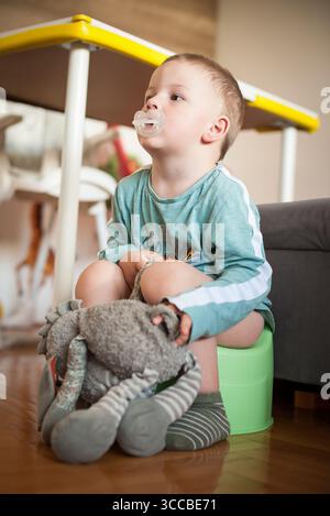 Ein Kleinkind mit Socken und einem langärmeligen Hemd sitzt auf einem Töpfchen mit einem Schnuller im Mund. Er hält ein Plüschtier in der Hand. Hintergrund inkl. Stockfoto