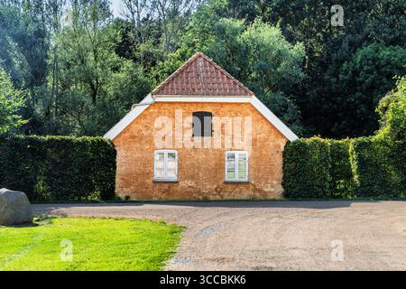 Ein malerisches Backsteinhaus ist von üppigen grünen Hecken umgeben, mit einem malerischen Wald, der im Hintergrund fasziniert und Charme, Gelassenheit und Landschaft weckt Stockfoto
