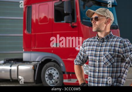 Ein LKW-Fahrer mit Kappe und Sonnenbrille posiert neben einem roten LKW und zeigt sein professionelles Verhalten auf einem Transportplatz unter freiem Himmel. Stockfoto