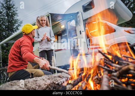 Ein Mann und ein Mädchen lachen und binden sich an einem Lagerfeuer vor ihrem Wohnmobil auf einem Campingplatz. Die Atmosphäre ist lebhaft, perfekt für Sommerabenteuer. Stockfoto