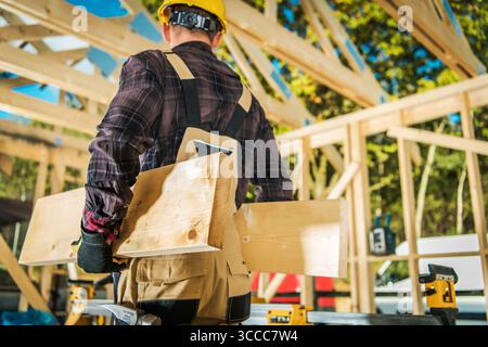 Ein Bauarbeiter mit Schutzhelm und Handschuhen transportiert Holzdielen auf eine sonnige Baustelle und zeigt die aktive Bauphase. Stockfoto