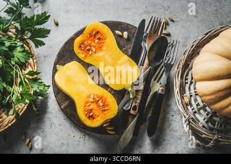 Halbierter Butternusskürbis auf Teller mit Besteck und Kräutern auf grauem Betonküchentisch Hintergrund. Zuhause kochen mit saisonalem Gemüse. Draufsicht. Stockfoto