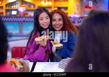 Zwei fröhliche Freundinnen genießen gemeinsam Pizzascheiben in einem lebhaften Restaurant und feiern ihre Freundschaft Stockfoto
