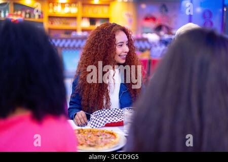 Glückliche junge Frau mit roten lockigen Haaren lächelt beim Abendessen mit Freunden in einer Pizzeria Stockfoto
