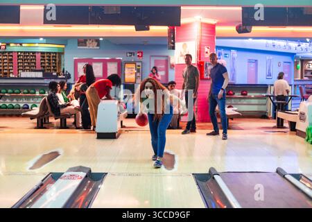 Fröhliche junge Frau wirft Bowlingball und genießt ein Spiel mit Freunden auf einer lebhaften Bowlingbahn Stockfoto