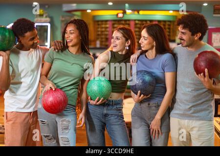Fünf junge Freunde spielen zusammen, halten ihre Bowlingbälle und lächeln Stockfoto