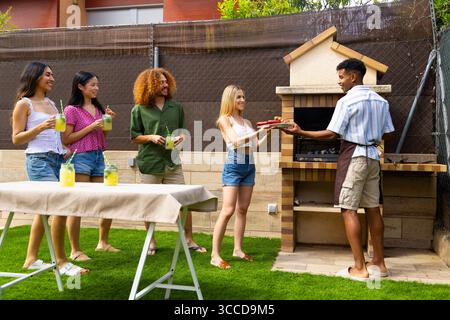 Gruppe von verschiedenen Freunden, die Spaß haben, Essen grillen und erfrischende Getränke bei einer Grillparty im Hinterhof trinken Stockfoto