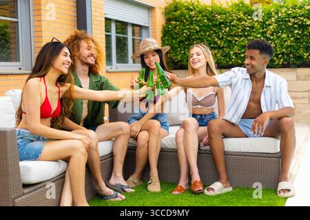 Gruppe fröhlicher junger, ethnischer Freunde, die während der Sommerparty auf dem Sofa im Garten mit Bierflaschen tosten Stockfoto