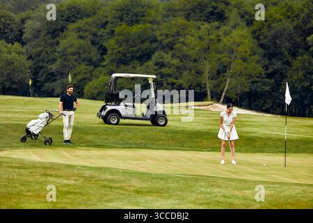 Ein Mann und eine Frau spielen gemeinsam auf einem wunderschön gepflegten, üppig grünen Golfplatz mit einem Golfwagen in der Nähe, der perfekt ist Stockfoto