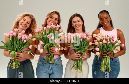 Gruppe von vier lächelnden Frauen verschiedener Ethnien, die frische rosa Tulpen halten. Freundschaft, Feier, Frühling, Schönheit, Glück, Vielfalt und Freude Stockfoto