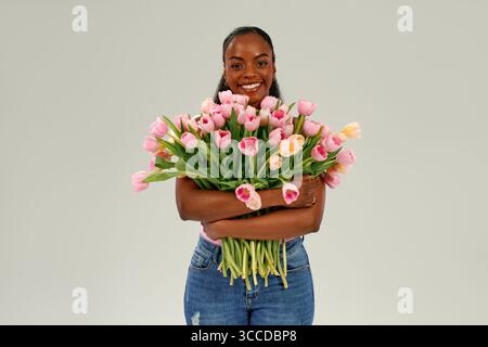 Afroamerikanische Frau, die rosa Tulpenstrauß hält, Mutter- oder Frauentag mit Freude und Liebe feiert, hell lächelnd auf neutralem Hintergrund. Konz Stockfoto