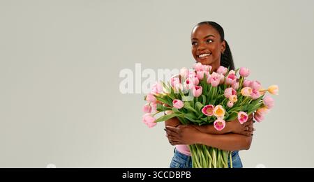 Afroamerikanische Frau, die rosa Tulpenstrauß hält, Mutter- oder Frauentag mit Freude und Liebe feiert, hell lächelnd auf neutralem Hintergrund. Konz Stockfoto