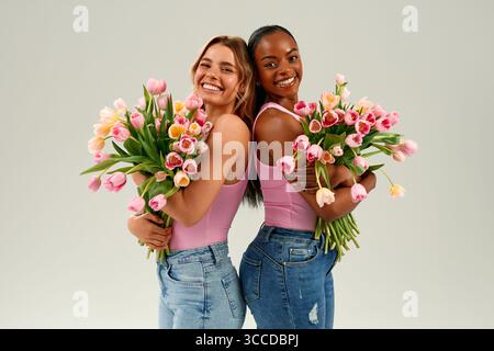 Zwei fröhliche Frauen, eine Kaukasierin, eine Afroamerikanerin, die frische Tulpensträuße hält, die hell auf weißem Hintergrund lächeln, perfekt für Muttertag oder Frau Stockfoto