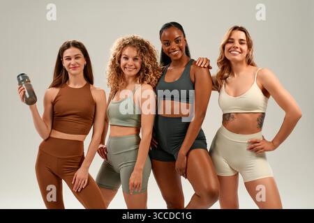 Vier verschiedene sportliche Frauen, darunter eine Afroamerikanerin, posieren gemeinsam in Sportkleidung und halten eine Wasserflasche auf weißem Hintergrund. Fitnes Stockfoto
