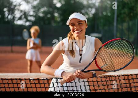 Die lächelnde junge Frau in Sportbekleidung lehnt sich auf das Tennisnetz und hält nach einem Spiel auf einem Tonplatz selbstbewusst Schläger. Das Konzept des Vertrauens, der Ermächtigung, A Stockfoto
