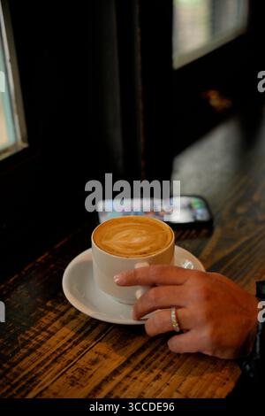 Frau genießt einen Cappuccino in einem Café, mit einem Smartphone auf dem Tisch neben der Tasse Stockfoto