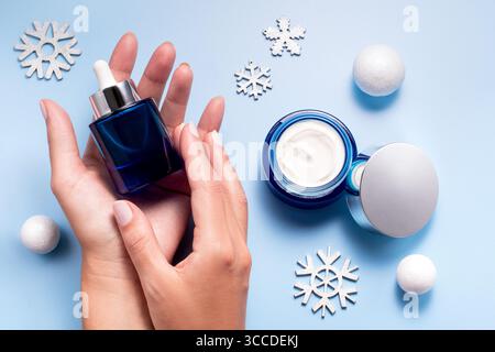 Schönheitskonzept der Winterpflege mit Frauenhänden, die Gesichtsserum und Creme auf blauem Hintergrund halten, Ansicht von oben Stockfoto