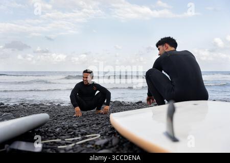 Zwei Surfer in Neoprenanzügen, die sich an einem Kiesstrand hocken und vor dem Surfen auf die Wellen des Ozeans schauen Stockfoto