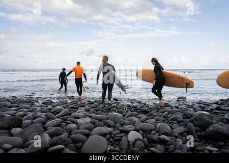 Gruppe von Surfern, die an einem schwarzen, felsigen Strand mit Surfbrettern spazieren gehen und das Meer für eine Surfsession betreten Stockfoto