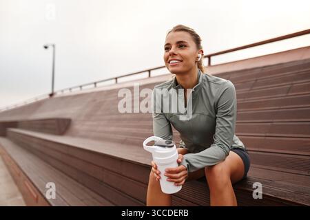 Eine junge Frau sitzt auf der Tribüne und lächelt hell, während sie nach Abschluss ihres Workouts eine Shaker-Flasche hält Stockfoto