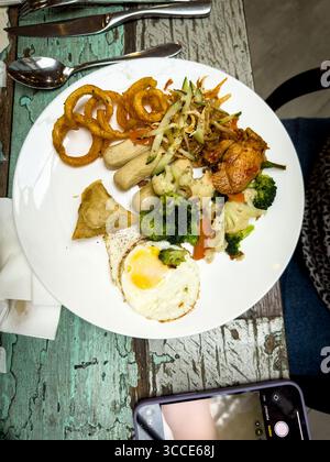 Flachbild-Lay gemischter Teller mit Spiegeleiern, Zwiebelringen, Samosa-Brokkoli-Gemüse auf rustikalem Tisch im Café-Restaurant kuala lumpur malaysia Stockfoto