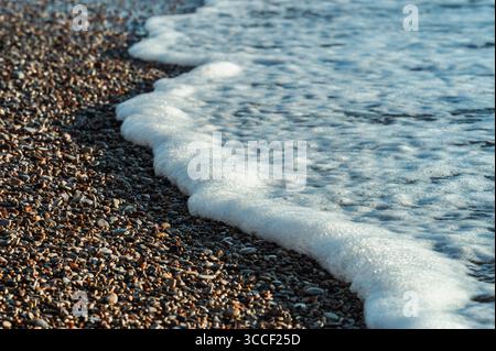 Weißer Meereswellen-Schaum, der über dem farbenfrohen Kieselstrand gespült wird. Natürliche Meereslandschaft, die den Treffpunkt des Meerwassers und der felsigen Küste mit sanfter Rundung zeigt Stockfoto