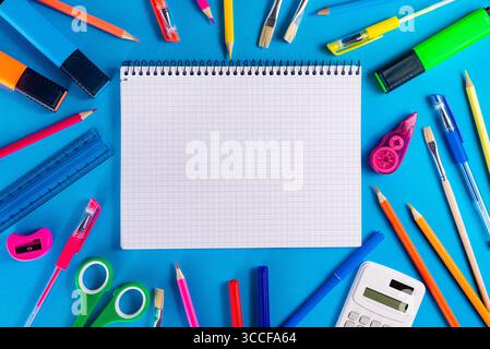 Flache Komposition verschiedener farbenfroher Schulpapiergegenstände, die um ein offenes quadratisches Notizbuch auf einem hellblauen Hintergrund angeordnet sind. Stockfoto