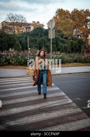 Attraktive Frau in braunem Mantel und Jeans, die auf Fußgängerüberquerung läuft Stockfoto