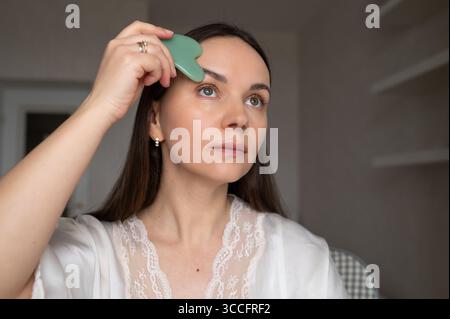 Junge Frau im Seidenmantel, die Gesichtsmassagen mit Gua Sha Schaber macht, während sie in den Spiegel blickt. Häusliche Hautpflege und ästhetische Verfahren Stockfoto