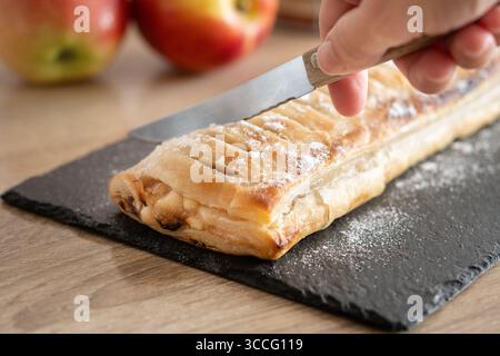 Nahaufnahme einer Person, die einen frisch gebackenen hausgemachten Apfelstrudel auf einem schwarzen Schieferbrett schneidet. Stockfoto