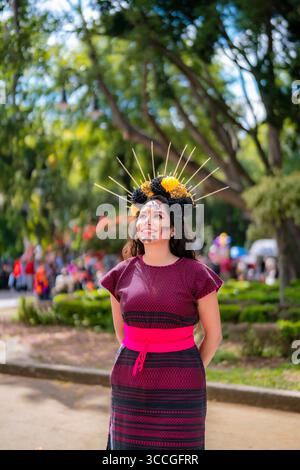 Die junge glückliche lateinfrau, die catrina Make-up und ein traditionelles gewebtes Kleid trägt, feiert Dia de Muertos im Central Park von Coyoacan. Ihre Blumenkrone, Co Stockfoto