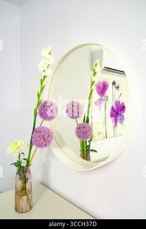 Stilvolle Inneneinrichtung mit einem lebendigen Blumenarrangement in einer rosafarbenen Vase auf einem weißen Konsolentisch, deutlich reflektiert in einem großen, kreisförmigen weiß-fra Stockfoto