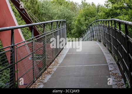 Eine moderne Metallbrücke mit Geländern kurvt durch üppiges grünes Holz unter einem bewölkten Himmel, die mit einer hohen, eckigen Millennium Bridge Calne verbunden ist Stockfoto