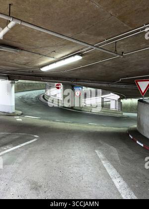 Gebogene Betonrampe in einem Flughafenparkhaus mit Richtungsschildern, Deckenbeleuchtung und lackierten Pfeilen, die den Verkehrsfluss und anzeigen Stockfoto