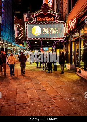 Fußgänger, die unter beleuchteten Schildern spazieren, darunter nationale Konzerte im Hard Rock Cafe am Times Square in New York City. Stockfoto